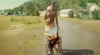 Photo still from the movie Crip Camp, with a camper in a wheelchair and camper standing behind them holding a guitar