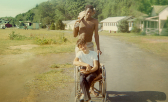 Photo still from the movie Crip Camp, with a camper in a wheelchair and camper standing behind them holding a guitar