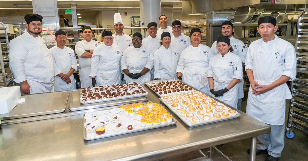 A group of culinary students and chefs in the kitchen next to desserts
